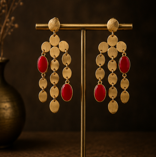 Gold-toned statement earrings with dangling oval and round gold discs and red teardrop beads, shown on a display stand, worn by a model in profile, and laid flat on a dark textured surface.