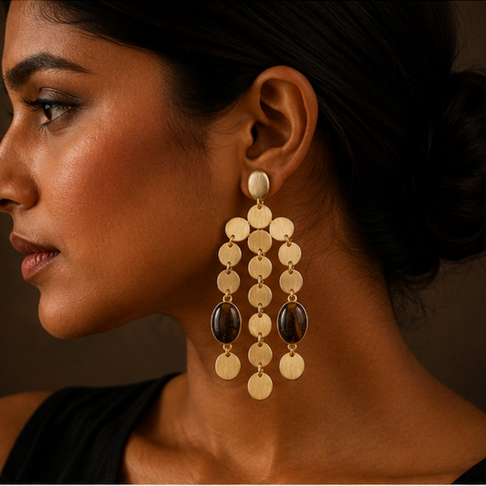 A close-up side profile of a woman wearing a large gold-toned dangle earring with circular disc charms and dark brown oval stones against a dark brown background.