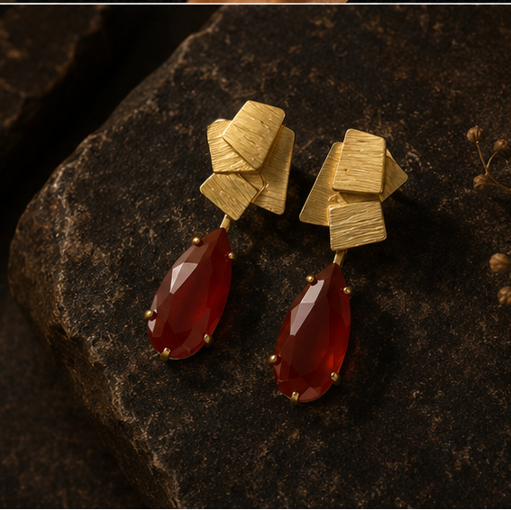 Collage showing a pair of statement earrings displayed on a gold stand, worn by a woman in a side-profile portrait, and resting on a dark textured surface. The earrings feature a textured gold geometric top with dangling teardrop-shaped deep red gemstones.